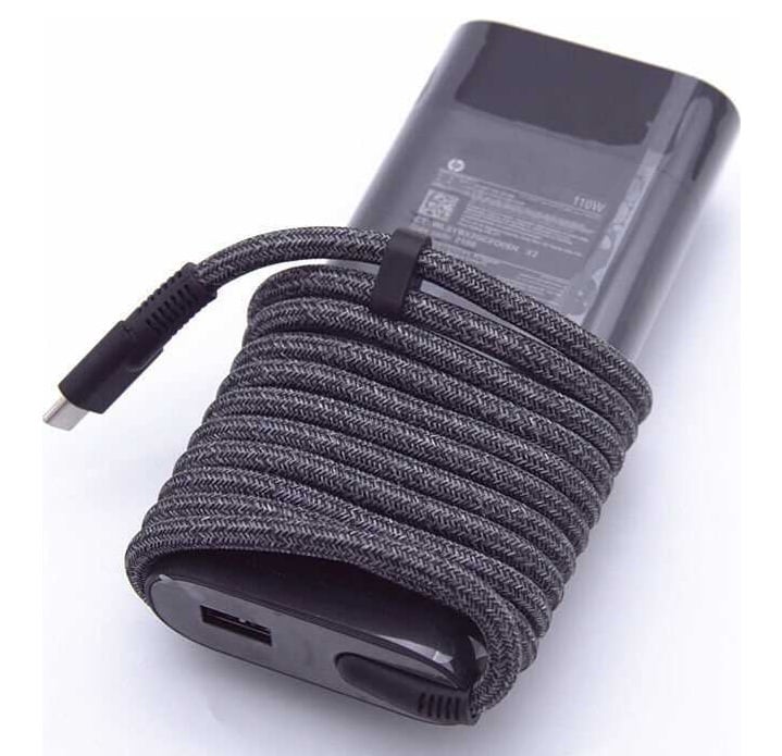 Angled perspective view of the black HP 110W USB-C AC power adapter (Model: M52950-001) showcasing its rectangular brick design wrapped neatly with a thick grey braided USB Type-C cable secured by a strap, alongside a visible auxiliary USB Type-A port on the front edge.